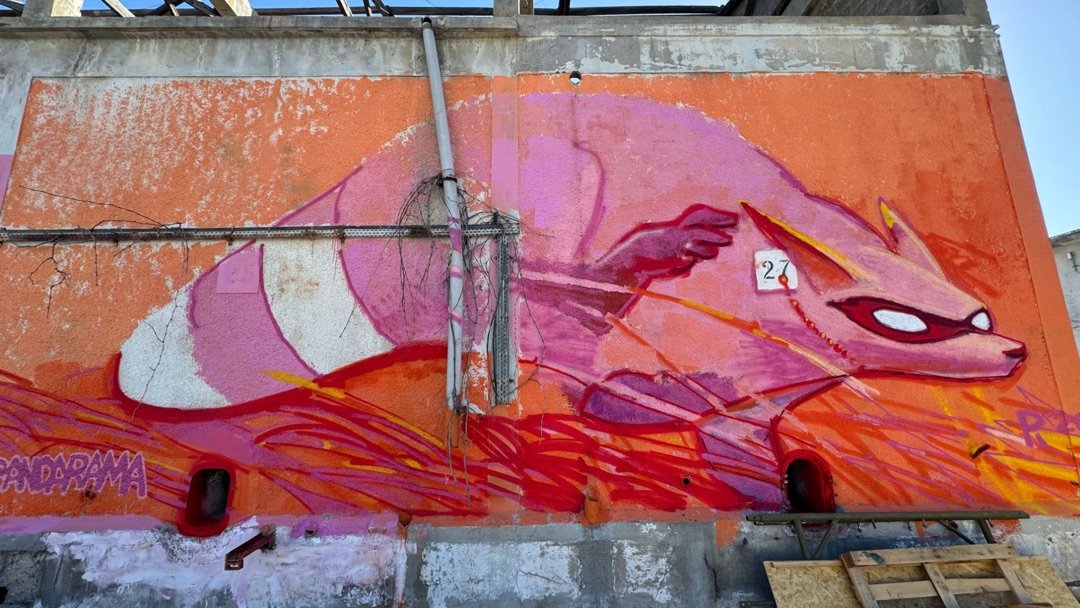 Pink masked creature with a long striped tail racing over red and orange streaks, painted across a weathered concrete wall